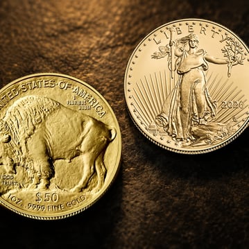Close-up of two American Gold bullion coins resting on a dark textured surface under warm golden lighting. A Gold Buffalo coin is shown in the foreground with the reverse design featuring the American bison, while an American Gold Eagle displays Lady Liberty walking confidently with a torch and olive branch. The shallow depth of field and rich lighting emphasize the coins' fine details and reflective gold finish.