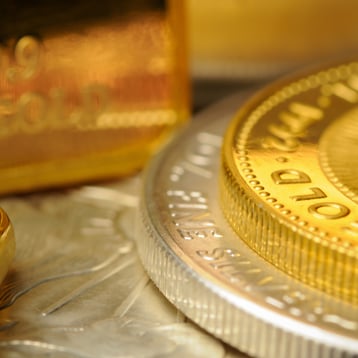Close-up image of stacked gold and silver bullion, with the edge of a gold coin in sharp focus and partial lettering visible, while surrounding coins and a gold bar appear softly blurred in the background under warm lighting.
