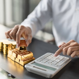 Person calculating gold bullion bar investment with stacked gold bars and coins on a desk