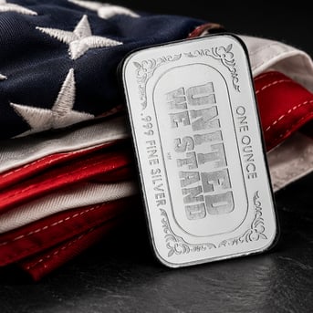 A Highland Mint one-ounce silver bar engraved with "United We Stand" leans against a neatly folded American flag on a dark stone surface. The close-up composition showcases the bar's detailed engraving and polished silver finish, while soft directional lighting creates a premium product photography look with patriotic colors in the background.