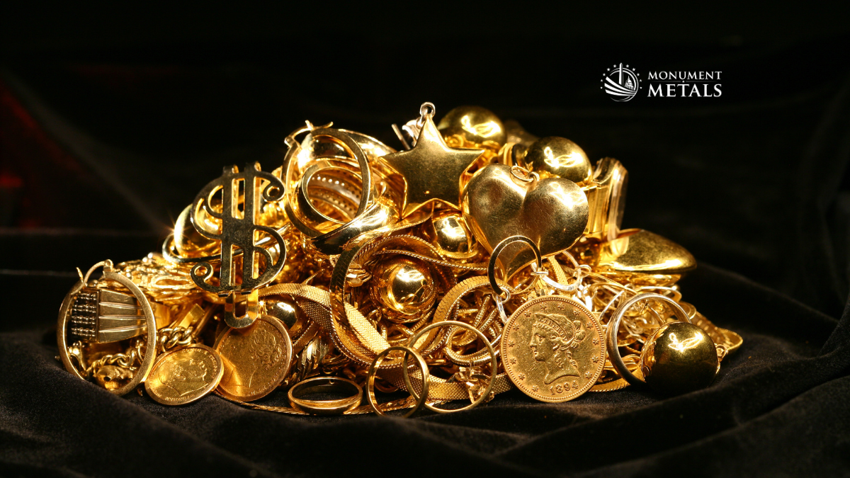 Pile of assorted gold jewelry and coins, including rings, chains, pendants, and vintage gold pieces, displayed on a dark fabric background with the Monument Metals logo in the upper right, representing inherited or scrap gold value.
