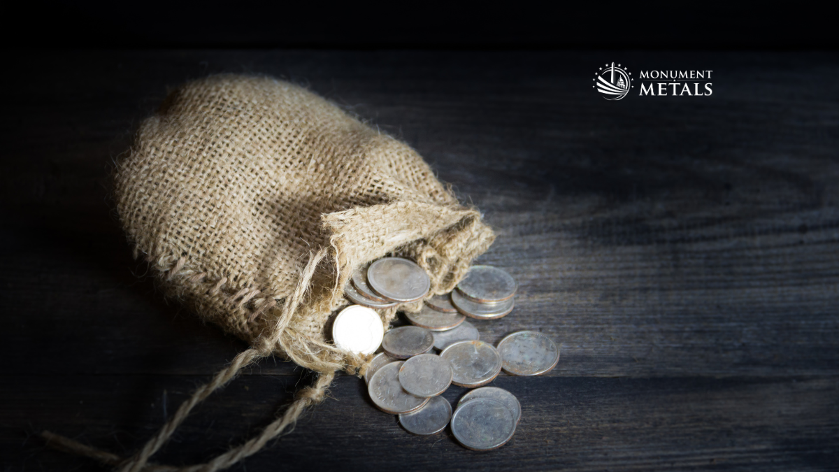 Burlap sack spilling pre-1965 junk silver coins including dimes and quarters on dark wood surface with Monument Metals logo