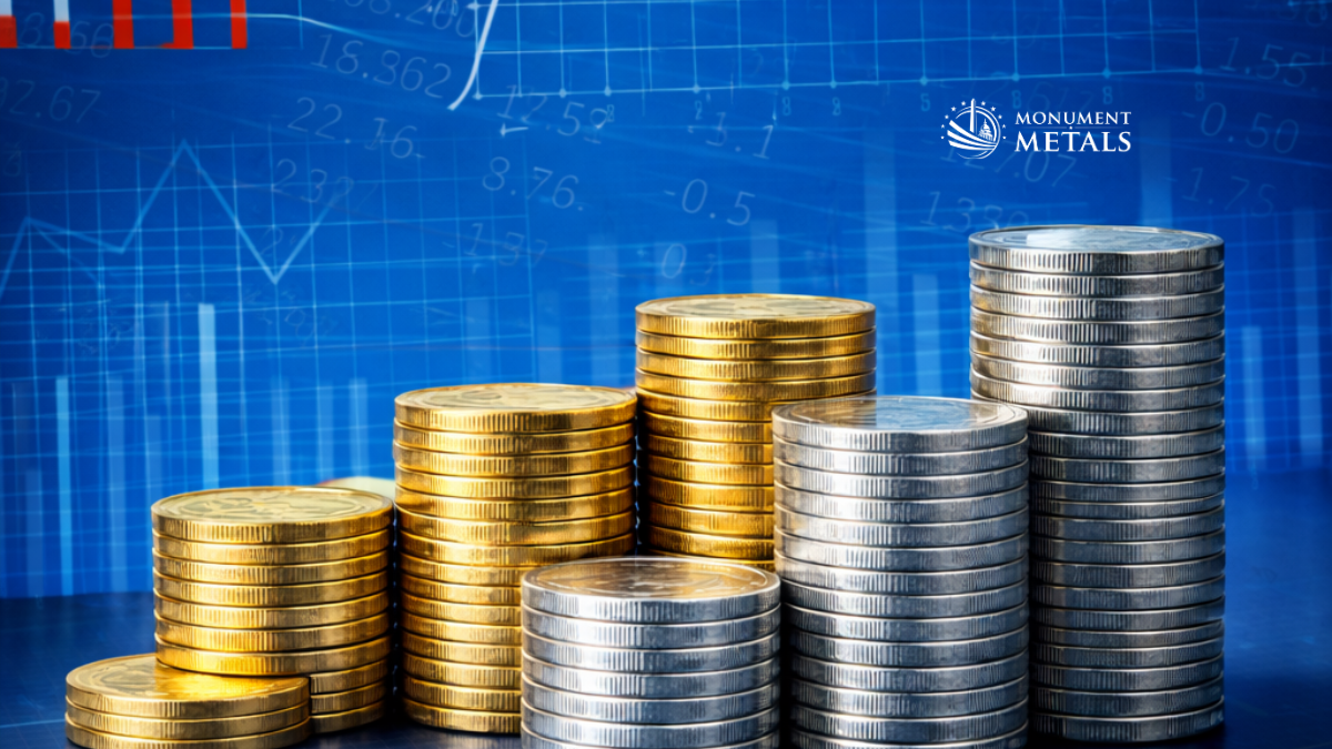 Stacks of gold and silver coins arranged in ascending order against a blue financial chart background, illustrating precious metals price movement and market trends, with Monument Metals logo.