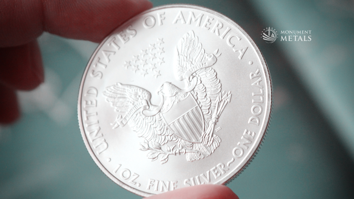 Close-up photograph of a 1 oz American Silver Eagle bullion coin being held between two fingers. The reverse design features a heraldic eagle with a shield, olive branch, arrows, and thirteen stars above. The coin's detailed engraving and frosted silver finish are sharply in focus, while the background is softly blurred in cool blue tones.