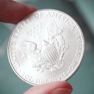 Close-up photograph of a 1 oz American Silver Eagle bullion coin being held between two fingers. The reverse design features a heraldic eagle with a shield, olive branch, arrows, and thirteen stars above. The coin's detailed engraving and frosted silver finish are sharply in focus, while the background is softly blurred in cool blue tones.