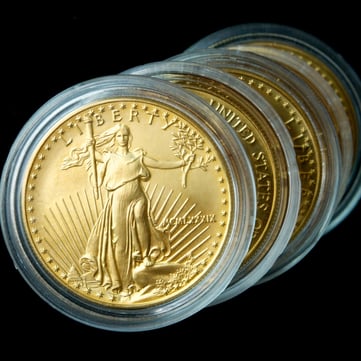 Close-up image of several American Gold Eagle bullion coins arranged in a staggered row against a black background. The foreground coin prominently displays Augustus Saint-Gaudens’ iconic Lady Liberty design, holding a torch and olive branch, with radiant sun rays behind her. Each gold coin is housed in a clear protective capsule, highlighting the rich golden luster and intricate details of the design.