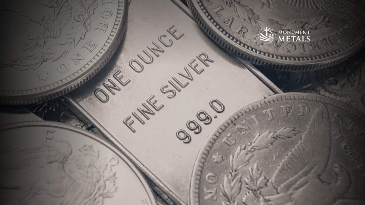 Close-up image of a one-ounce .999 fine silver bar surrounded by classic silver coins, showing detailed engravings and textures, with the Monument Metals logo in the upper right.