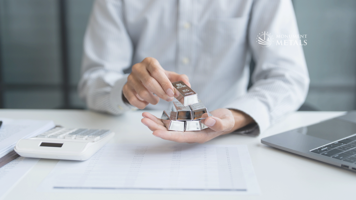 Person holding stacked silver bars in one hand while reviewing financial documents at a desk with a calculator and laptop