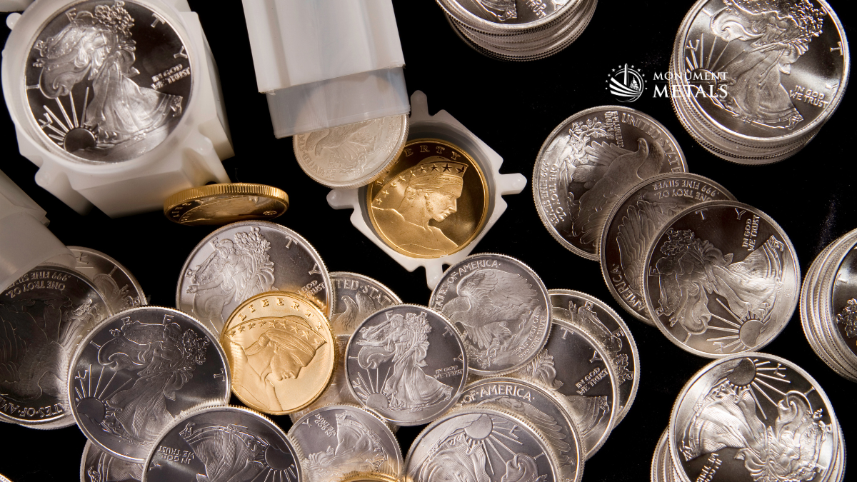 Close-up of gold and silver bullion coins including American Silver Eagles and gold coins, some stacked and some in protective tubes, displayed on a dark background with Monument Metals logo visible