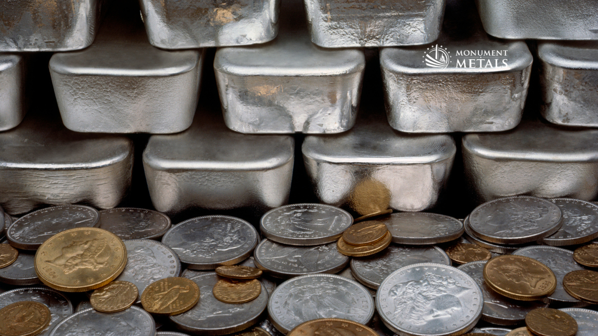 Stacked silver bullion bars with a spread of gold and silver coins in the foreground, illustrating the relationship between gold and silver values, with Monument Metals logo.