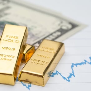 Assorted gold bars displayed over U.S. hundred-dollar bills and financial documents.